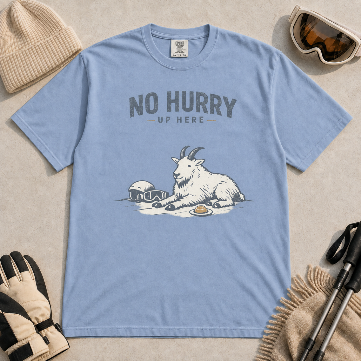 Washed denim blue t-shirt featuring a mountain goat relaxing in the snow with a helmet, goggles and snack beneath the words “No Hurry Up Here.”