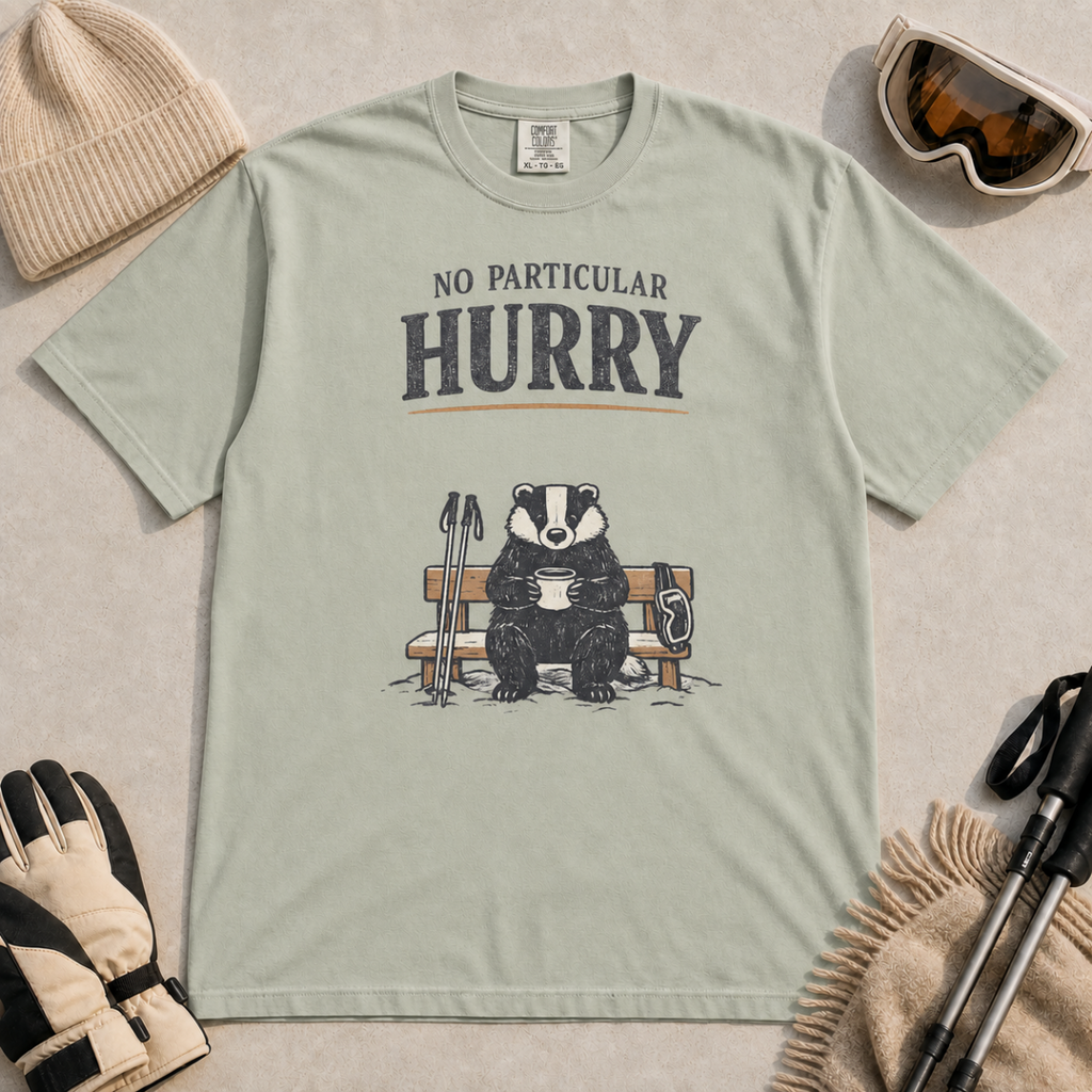 Bay green t-shirt featuring a badger relaxing on a snowy bench with a mug, ski poles and goggles beneath the words “No Particular Hurry.”