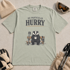 Bay green t-shirt featuring a badger relaxing on a snowy bench with a mug, ski poles and goggles beneath the words “No Particular Hurry.”