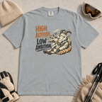 Granite gray t-shirt featuring a relaxed mountain goat holding a mug beside the phrase “High Altitude, Low Ambition.”