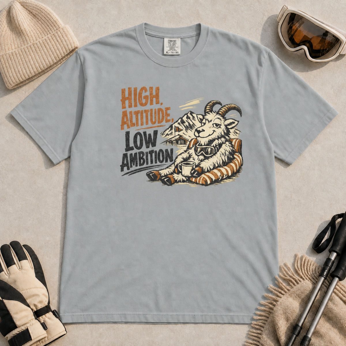 Granite gray t-shirt featuring a relaxed mountain goat holding a mug beside the phrase “High Altitude, Low Ambition.”