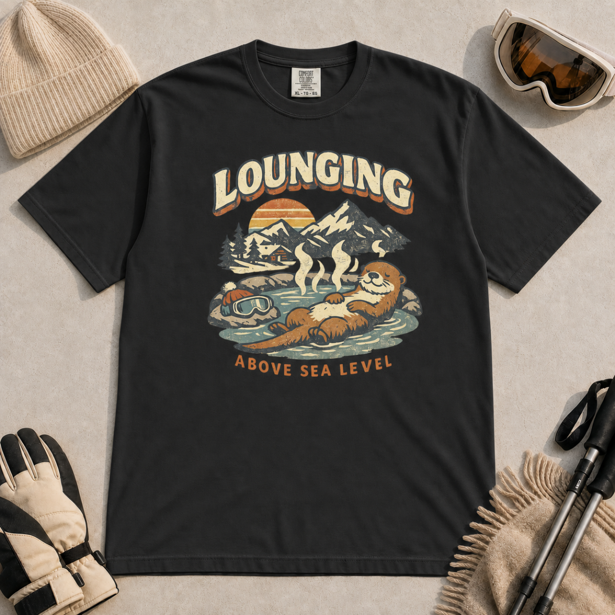 Black t-shirt featuring an otter relaxing in a steaming mountain hot spring beneath the words “Lounging Above Sea Level.”