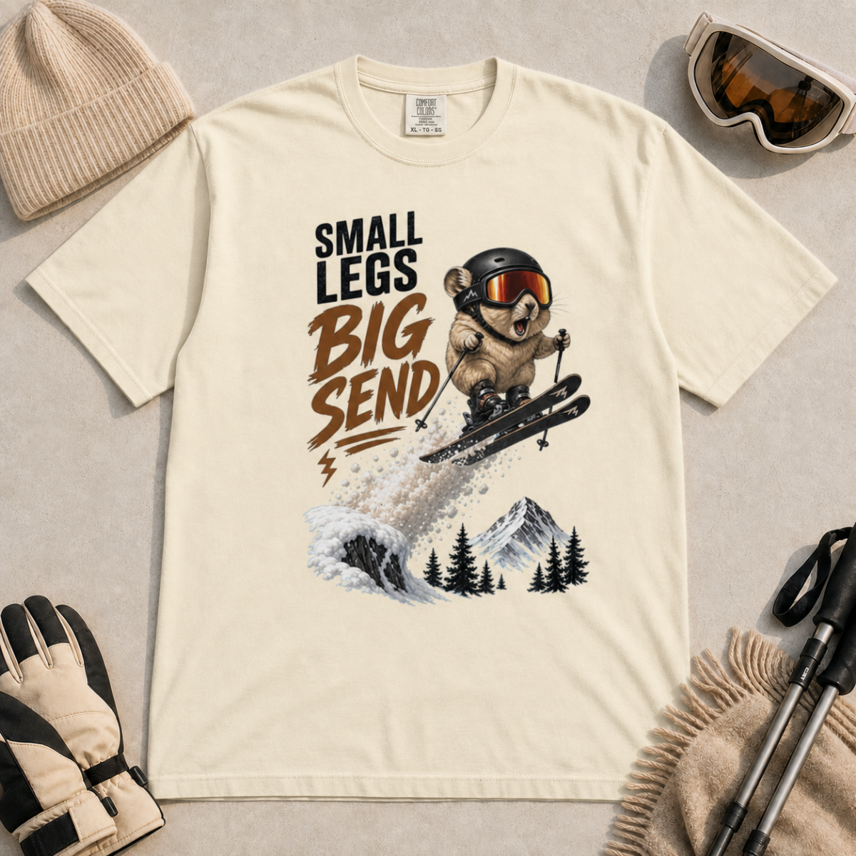 Ivory white t-shirt featuring a helmeted pika launching off a snowy ski jump with the phrase “Small Legs Big Send.”