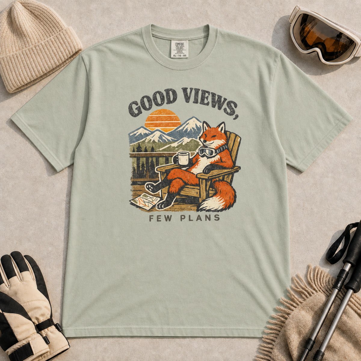 Bay green t-shirt featuring a relaxed fox in a wooden chair holding a mug against a mountain sunset with the phrase “Good Views, Few Plans.”