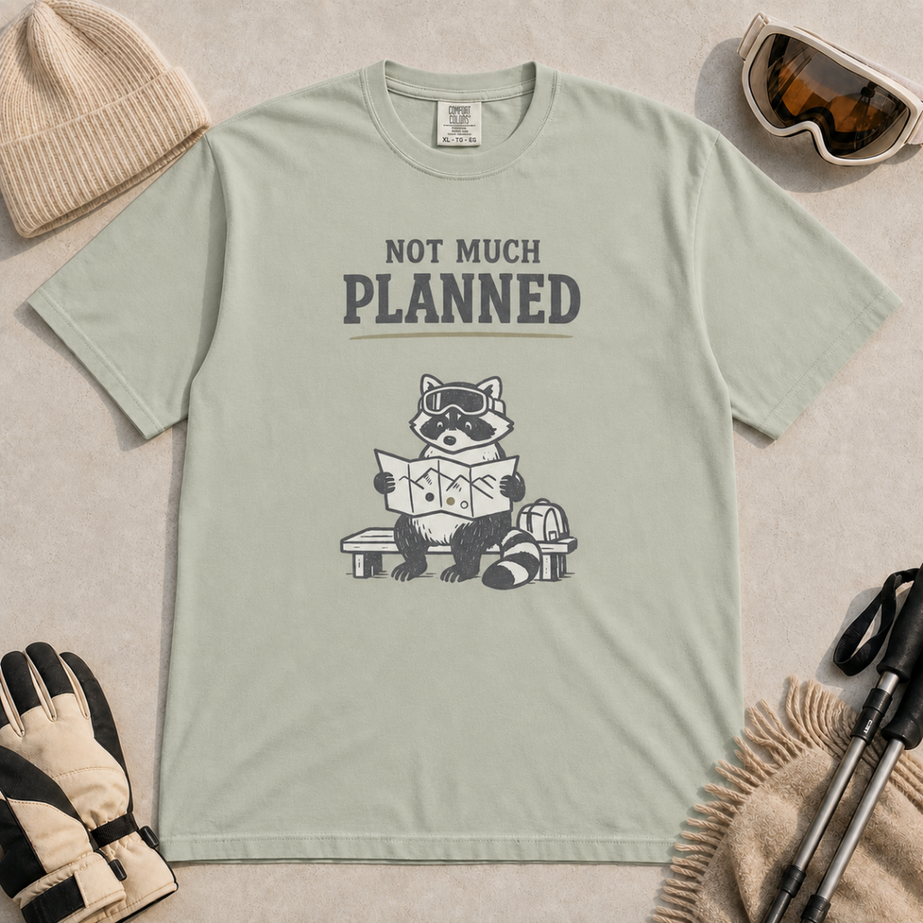 Khaki t-shirt featuring a raccoon in ski goggles sitting on a bench and studying a trail map, with the words “Not Much Planned.”