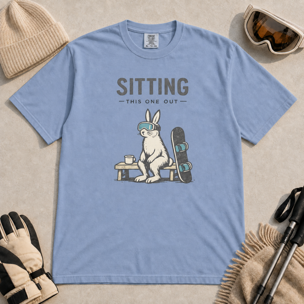 Washed denim blue t-shirt featuring a goggle-wearing rabbit resting on a bench beside a mug and snowboard with “Sitting This One Out” text.
