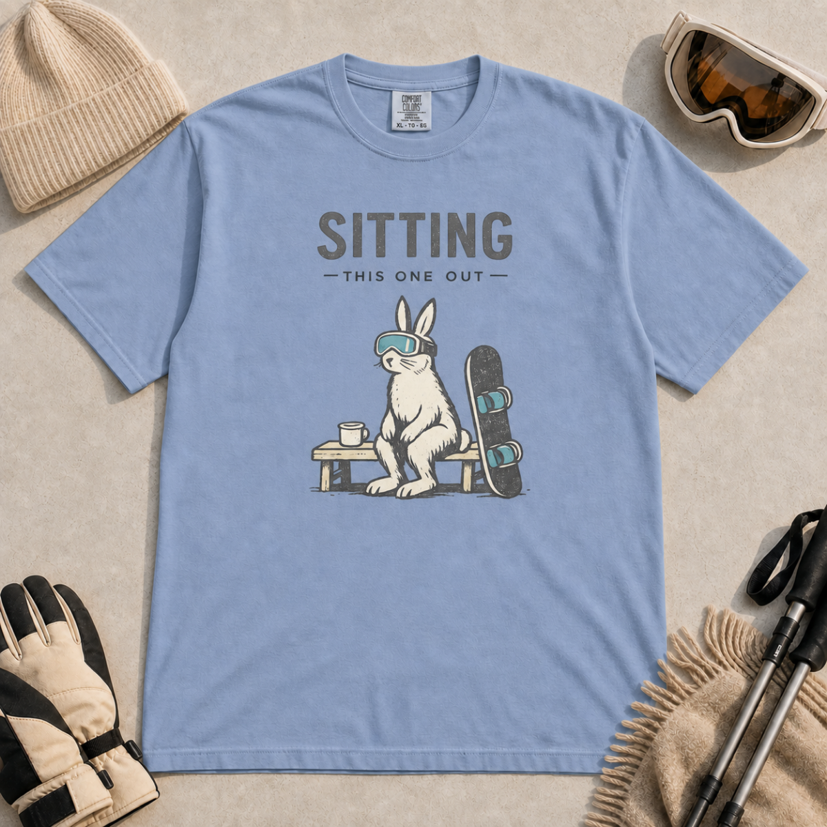 Washed denim blue t-shirt featuring a goggle-wearing rabbit resting on a bench beside a mug and snowboard with “Sitting This One Out” text.
