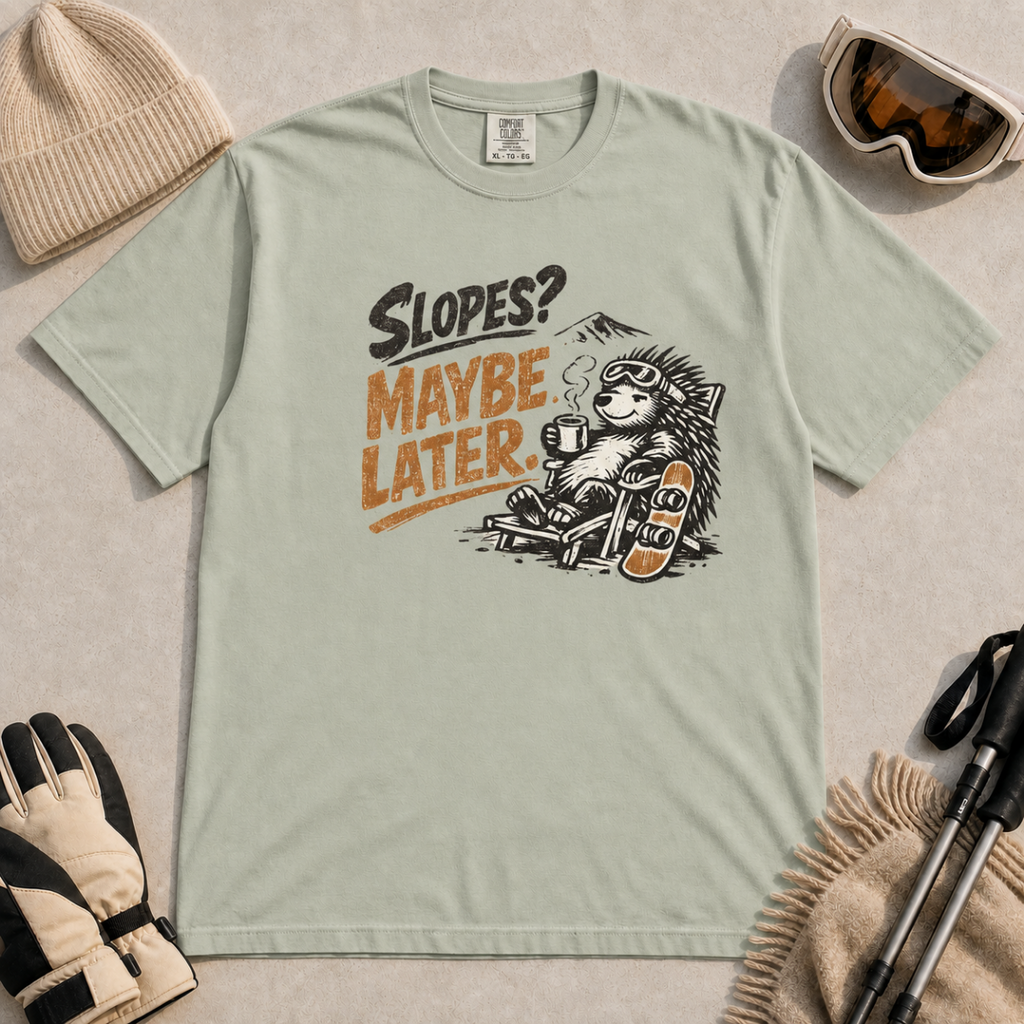 Bay green t-shirt featuring a relaxed hedgehog in a chair with a hot drink and snowboard beneath “Slopes? Maybe Later.” text.