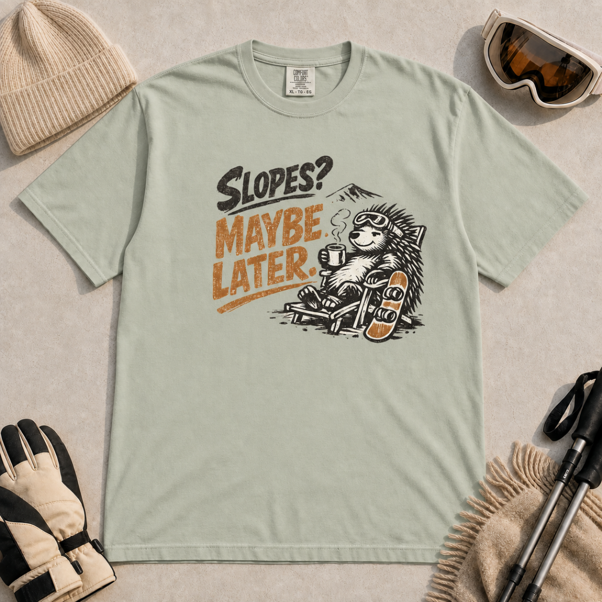 Bay green t-shirt featuring a relaxed hedgehog in a chair with a hot drink and snowboard beneath “Slopes? Maybe Later.” text.