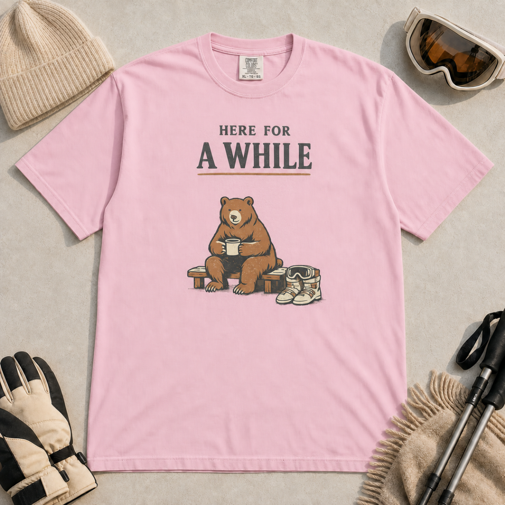 Blossom pink t-shirt featuring a seated bear holding a mug beside ski boots and goggles with the phrase “Here for a While.”