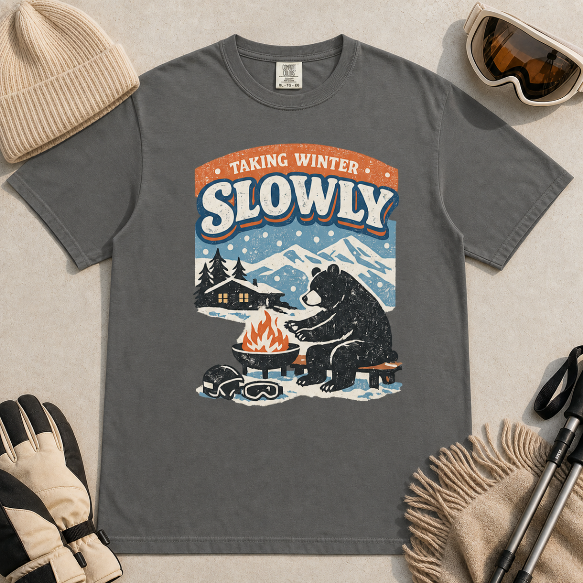 Pepper gray t-shirt featuring a bear warming its paws by a fire beneath “Taking Winter Slowly” text.