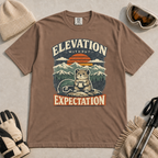 Espresso brown t-shirt featuring a mountain rodent with a mug and ski goggles beneath retro sunset scenery and the phrase “Elevation Without Expectation.”