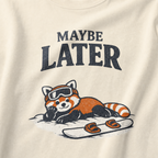 Close-up of an ivory white t-shirt design featuring a red panda resting in the snow beside a snowboard beneath the words “Maybe Later.”