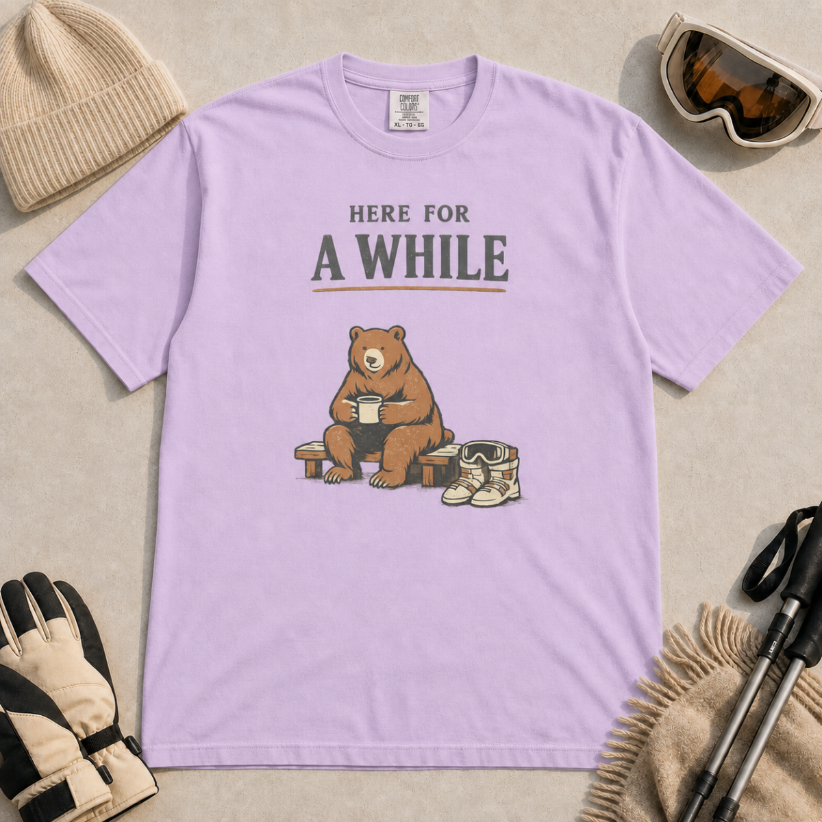 Orchid purple bear holding a mug beside ski boots and goggles with the phrase “Here for a While.”