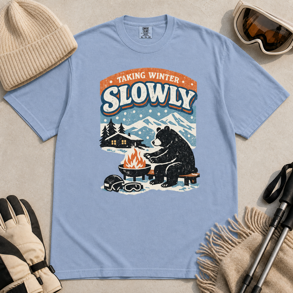 Washed denim blue blue t-shirt featuring a bear warming its paws by a fire beneath “Taking Winter Slowly” text.