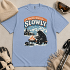 Washed denim blue blue t-shirt featuring a bear warming its paws by a fire beneath “Taking Winter Slowly” text.