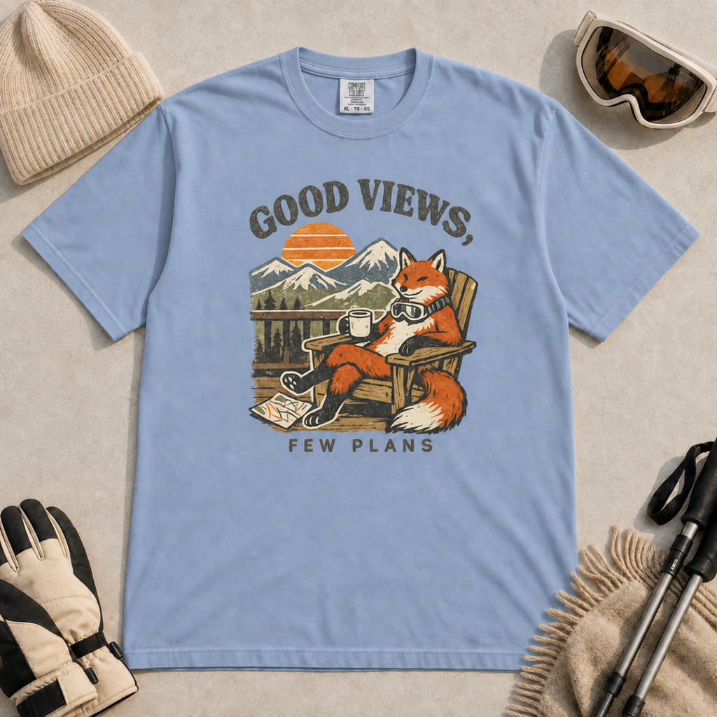 Washed denim blue t-shirt featuring a relaxed fox in a wooden chair holding a mug against a mountain sunset with the phrase “Good Views, Few Plans.”