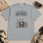 Granite gray t-shirt featuring a badger relaxing on a snowy bench with a mug, ski poles and goggles beneath the words “No Particular Hurry.”