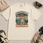 Ivory white t-shirt featuring a mountain rodent with a mug and ski goggles beneath retro sunset scenery and the phrase “Elevation Without Expectation.”