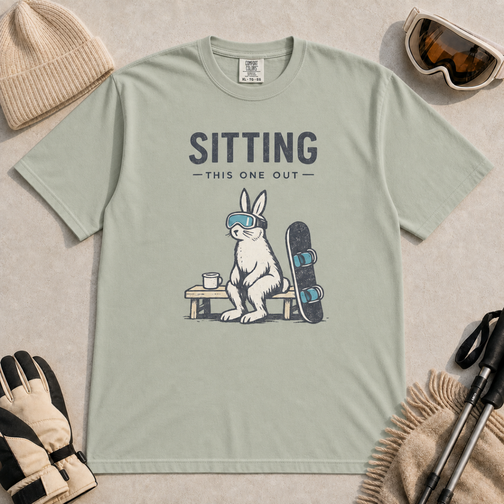 Bay green t-shirt featuring a goggle-wearing rabbit resting on a bench beside a mug and snowboard with “Sitting This One Out” text.