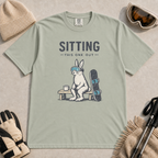 Bay green t-shirt featuring a goggle-wearing rabbit resting on a bench beside a mug and snowboard with “Sitting This One Out” text.