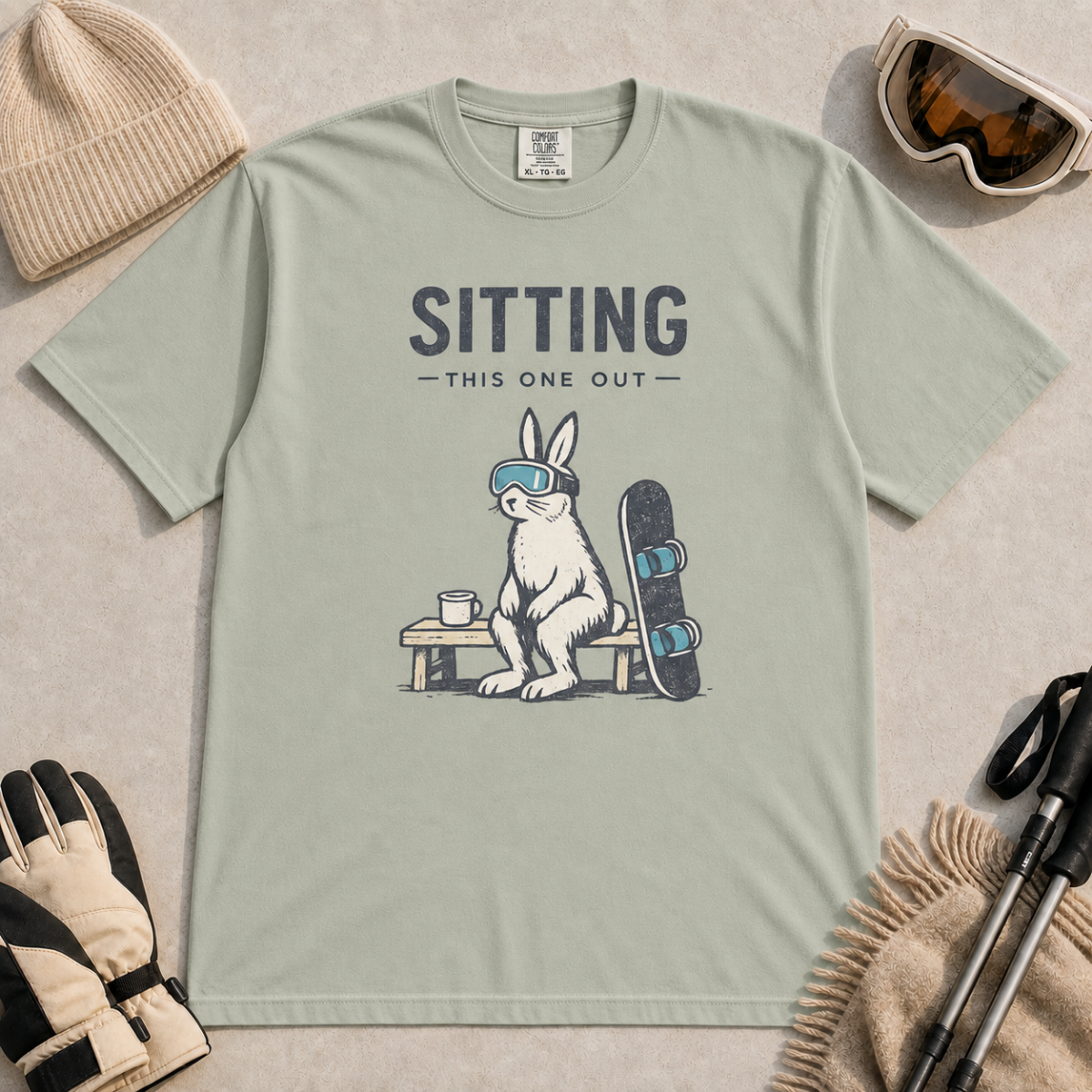 Bay green t-shirt featuring a goggle-wearing rabbit resting on a bench beside a mug and snowboard with “Sitting This One Out” text.