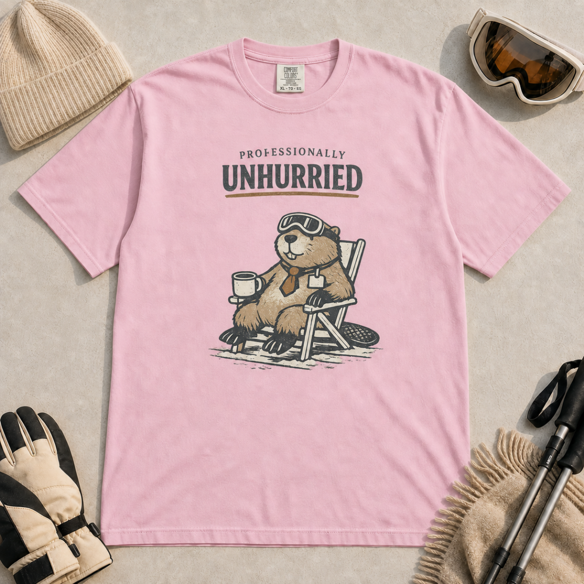 Blossom pink t-shirt featuring a relaxed beaver in ski goggles and a necktie, seated with a mug beneath “Professionally Unhurried” lettering.