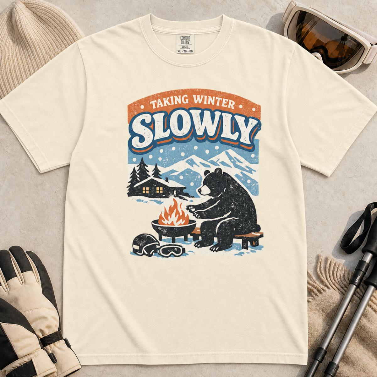 Ivory white t-shirt featuring a bear warming its paws by a fire beneath “Taking Winter Slowly” text.