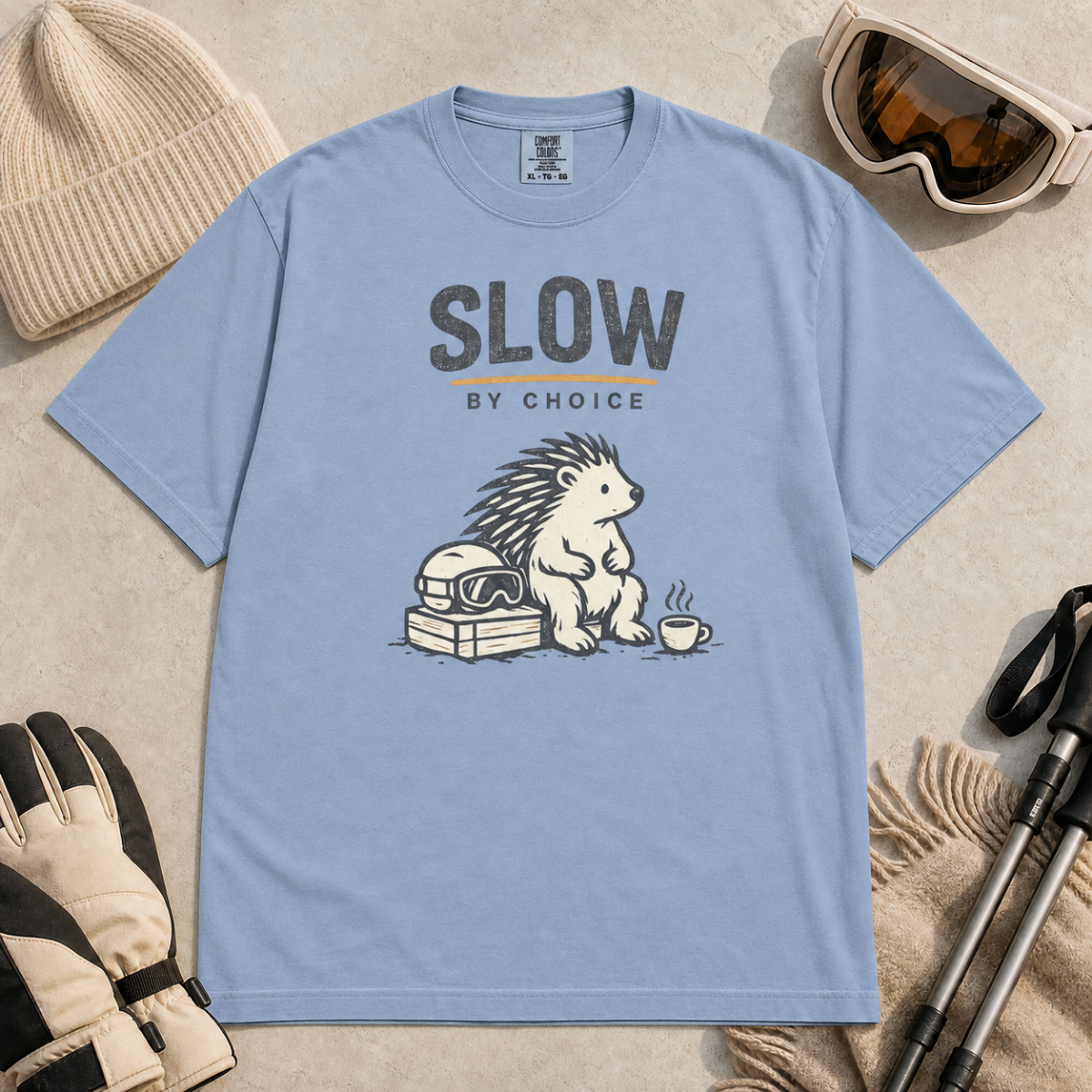 Washed denim blue t-shirt featuring a seated hedgehog beside ski goggles and a hot drink beneath “Slow by Choice” text.