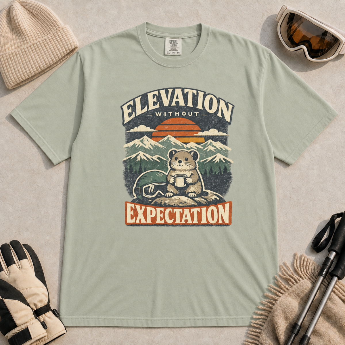 Bay green t-shirt featuring a mountain rodent with a mug and ski goggles beneath retro sunset scenery and the phrase “Elevation Without Expectation.”