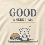 Close-up of an ivory white t-shirt featuring a small white bear holding a warm drink beside ski goggles with the phrase “Good Where I Am.”