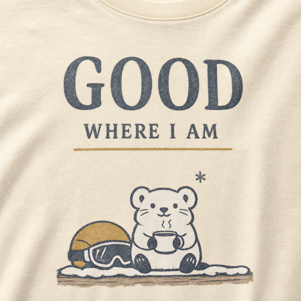 Close-up of an ivory white t-shirt featuring a small white bear holding a warm drink beside ski goggles with the phrase “Good Where I Am.”