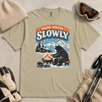 Khaki t-shirt featuring a bear warming its paws by a fire beneath “Taking Winter Slowly” text.