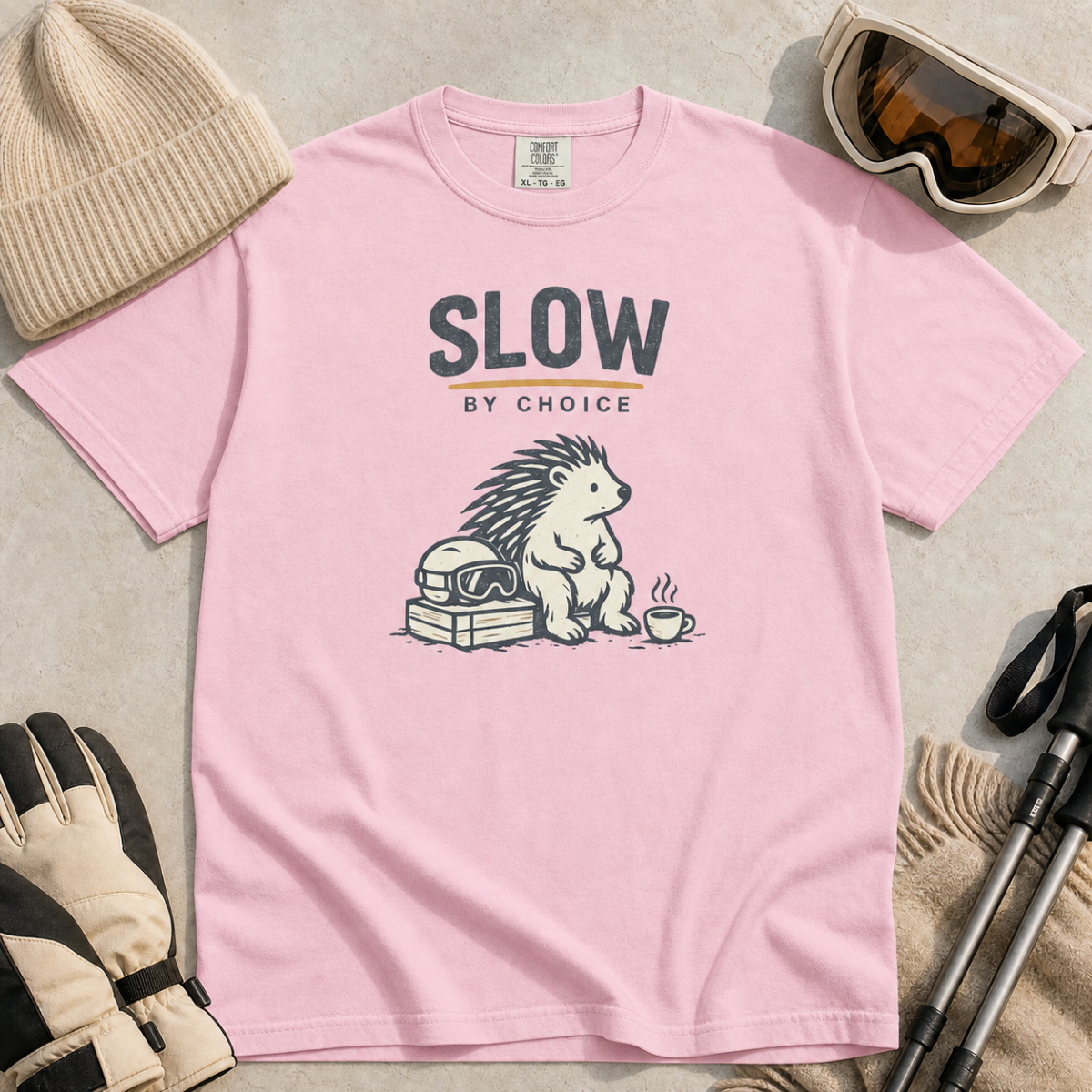 Blossom pink t-shirt featuring a seated hedgehog beside ski goggles and a hot drink beneath “Slow by Choice” text.