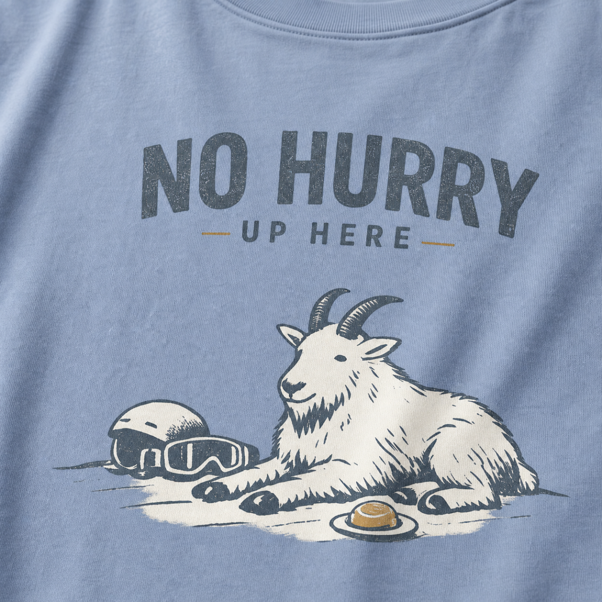 Close-up of a washed denim blue t-shirt featuring a mountain goat resting in the snow beside a helmet, goggles and snack beneath the words “No Hurry Up Here.”