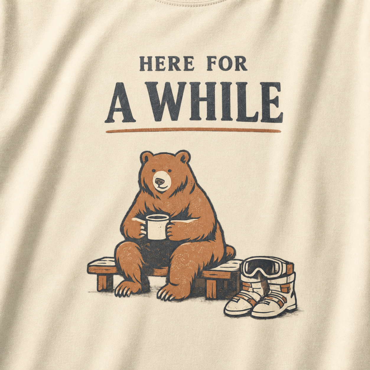 Close-up of an ivory white t-shirt featuring a bear holding a mug beside ski boots and goggles with the phrase “Here for a While.”