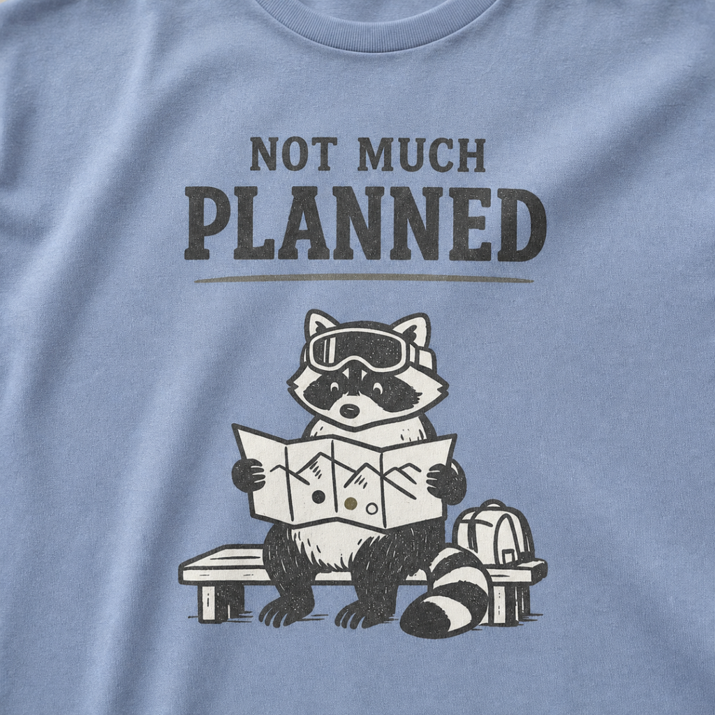 Close-up of a washed denim blue t-shirt featuring a raccoon in ski goggles holding a mountain map beside a backpack, with the words “Not Much Planned.”
