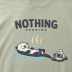Close-up of a bay green t-shirt featuring a relaxed otter floating in a hot spring beside ski goggles and a beanie, with the words “Nothing Pending.”