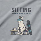 Close-up of a rabbit in ski goggles sitting beside a mug and snowboard beneath “Sitting This One Out” lettering.