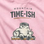 Close-up of a blossom pink t-shirt design showing a relaxed marmot with ski goggles, a clock and steaming mug beneath the words “Mountain Time-ish.”