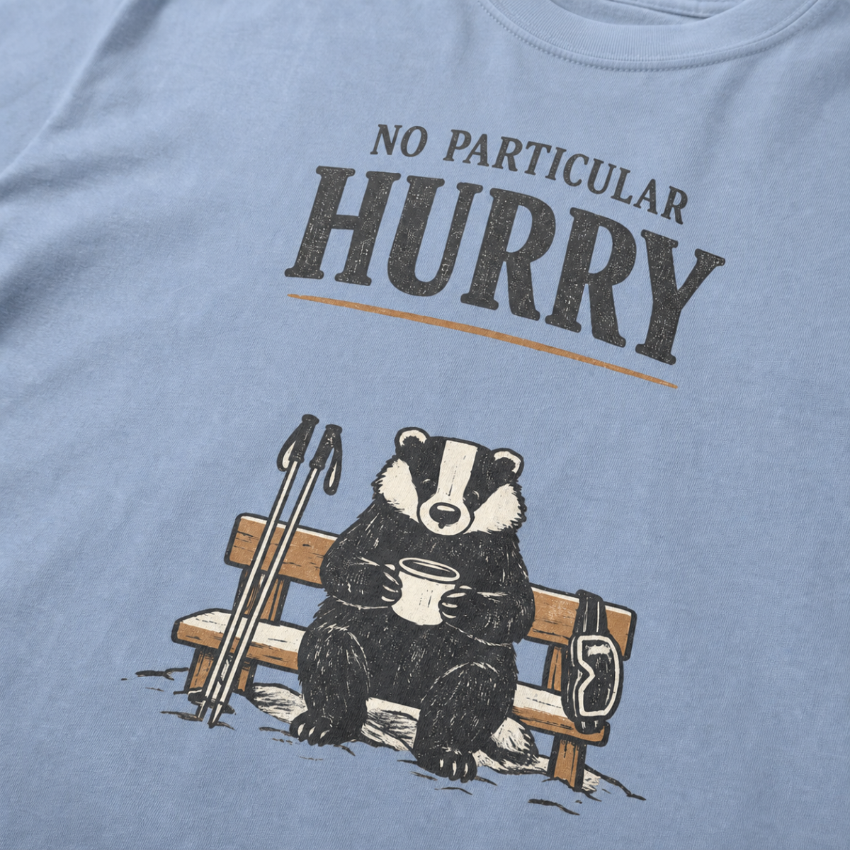 Close-up of a washed denim blue t-shirt featuring a badger seated on a snowy bench with a mug, ski poles and goggles beneath the words “No Particular Hurry.”