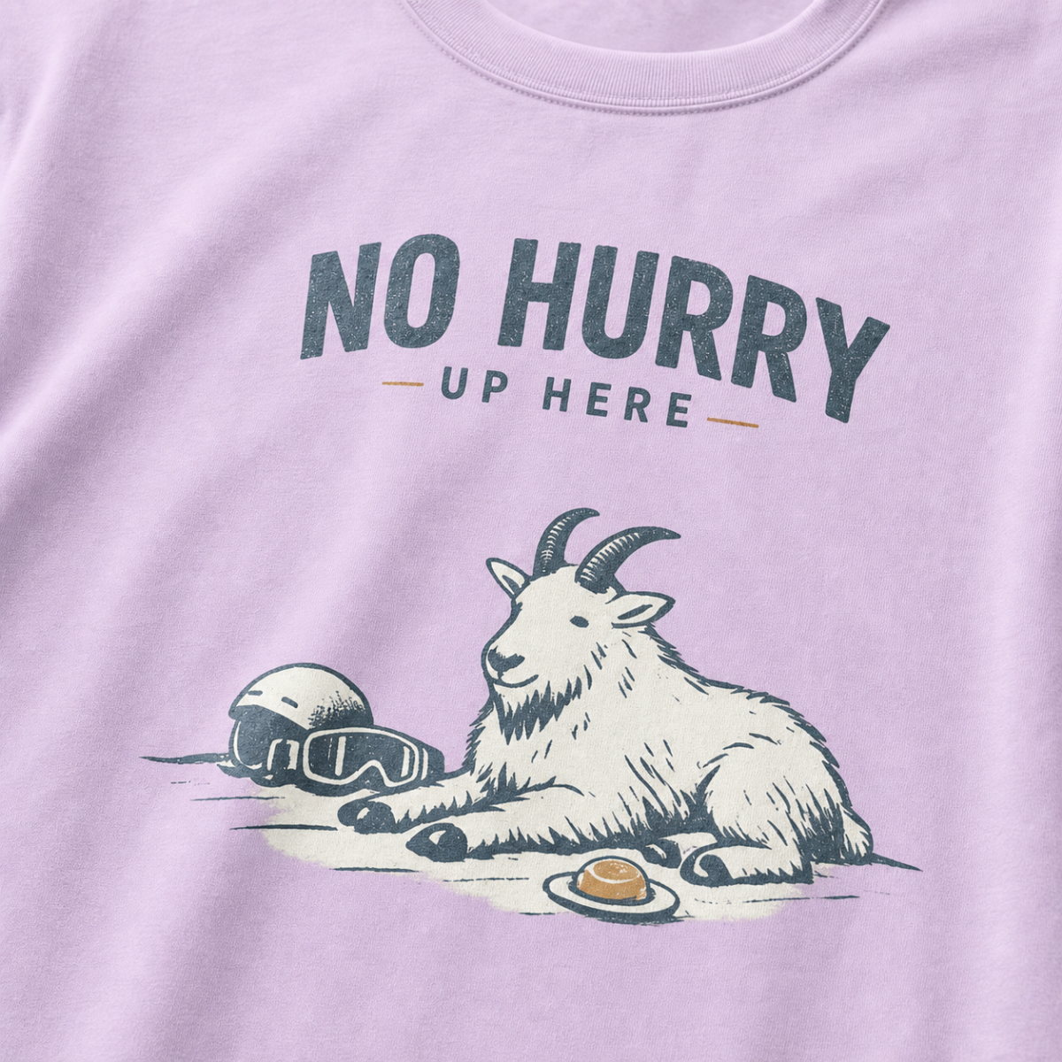 Close-up of a orchid purple t-shirt featuring a mountain goat resting in the snow beside a helmet, goggles and snack beneath the words “No Hurry Up Here.”