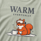 Close-up of a bay green t-shirt design showing a ski-goggle-wearing squirrel beside a hot drink under “Warm Eventually” text.