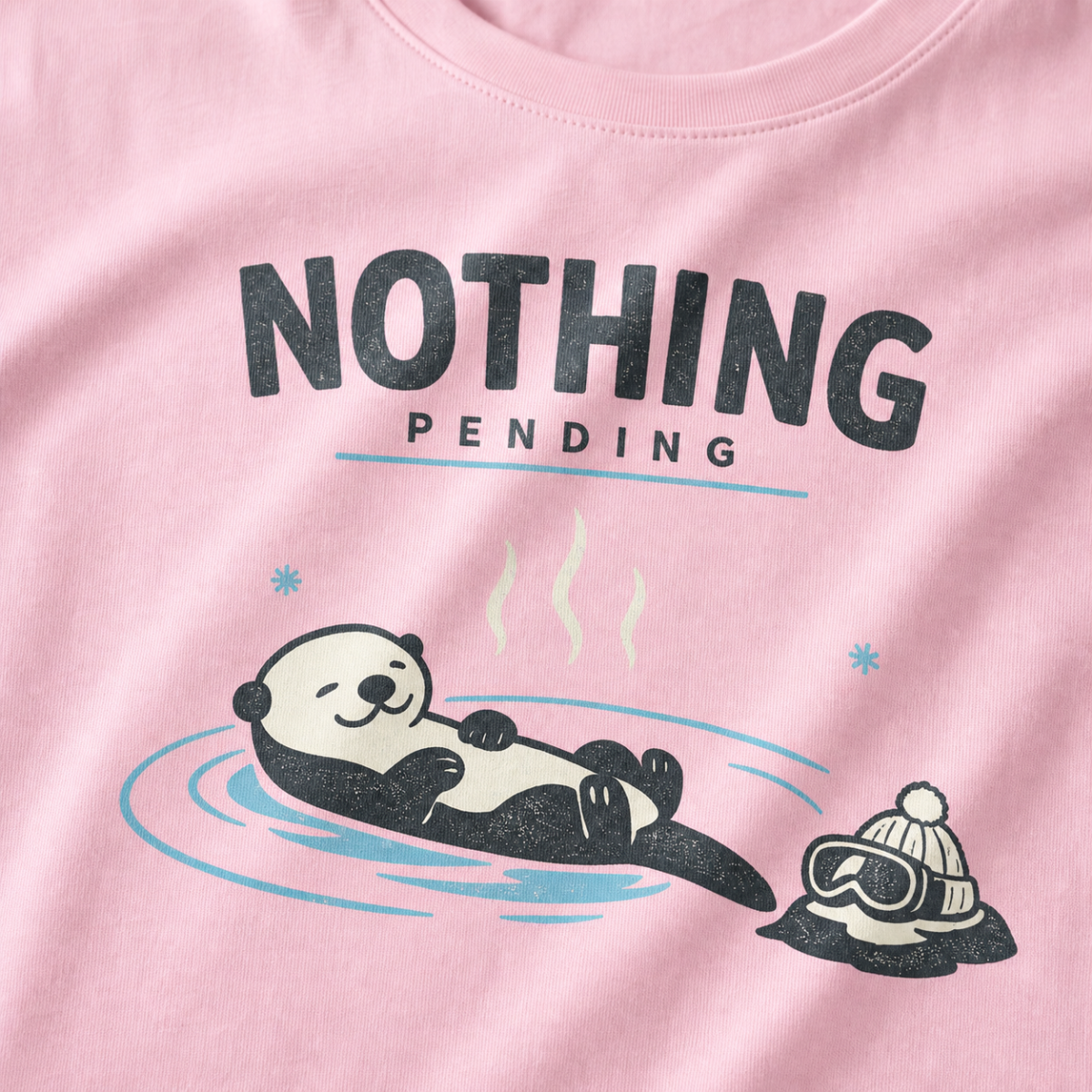 Close-up of a blossom pink t-shirt featuring a relaxed otter floating in a hot spring beside ski goggles and a beanie, with the words “Nothing Pending.”
