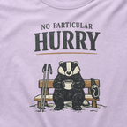 Close-up of an orchid purple t-shirt featuring a badger seated on a snowy bench with a mug, ski poles and goggles beneath the words “No Particular Hurry.”