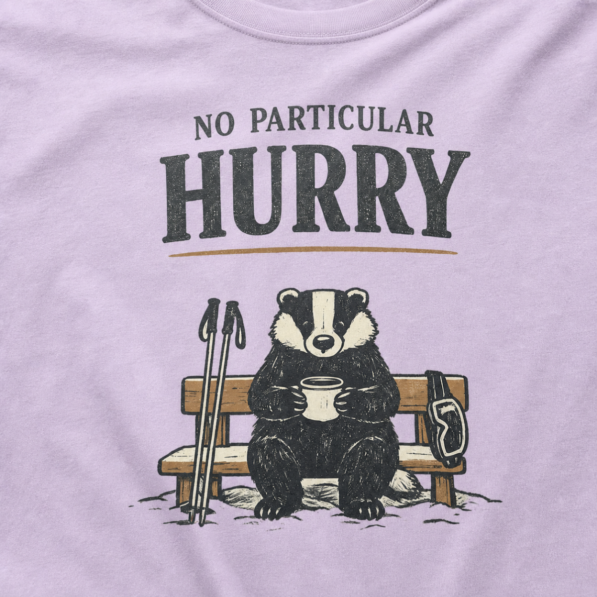Close-up of an orchid purple t-shirt featuring a badger seated on a snowy bench with a mug, ski poles and goggles beneath the words “No Particular Hurry.”