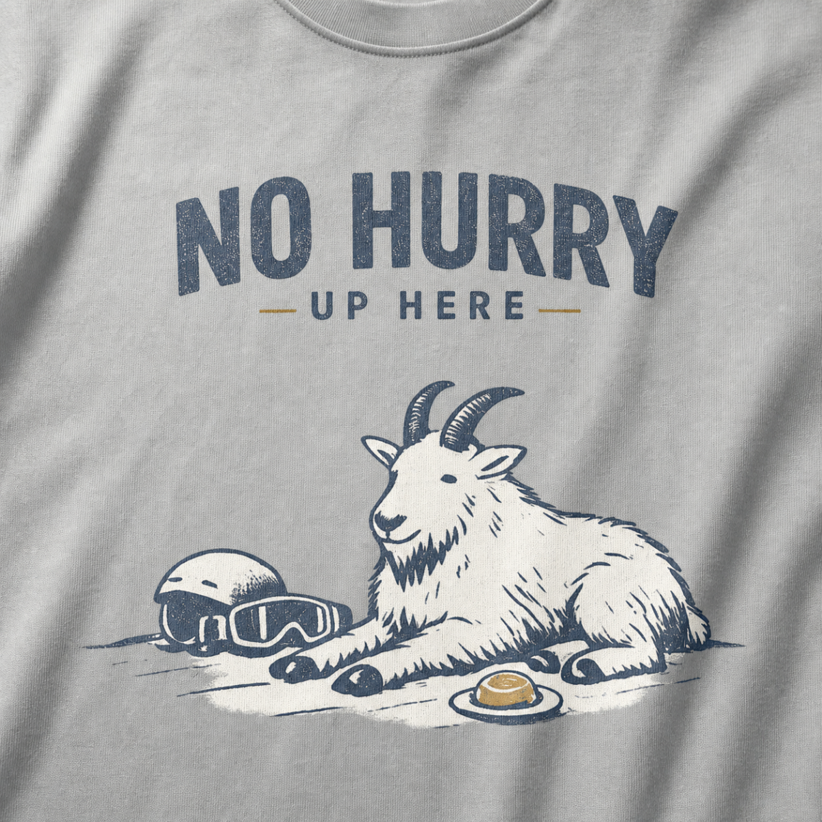 Close-up of a granite gray t-shirt featuring a mountain goat resting in the snow beside a helmet, goggles and snack beneath the words “No Hurry Up Here.”