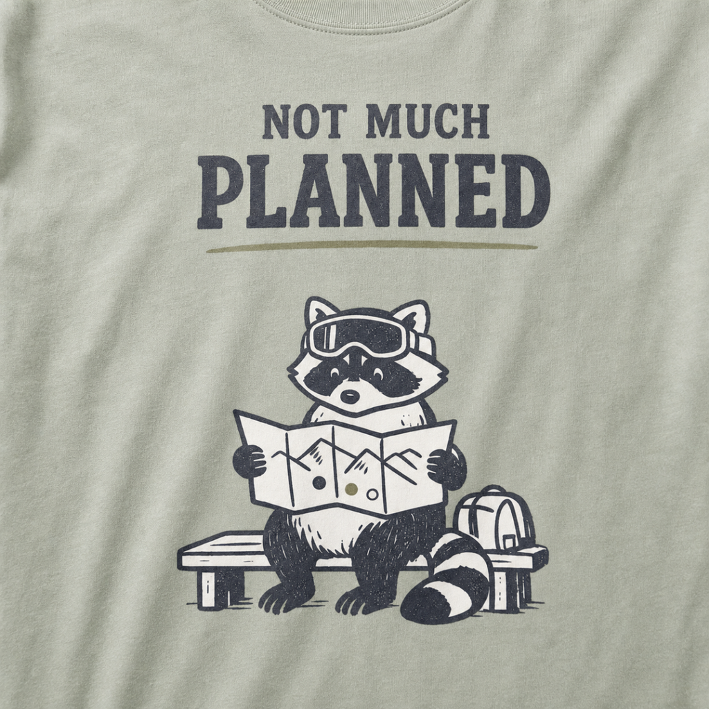 Close-up of a khaki t-shirt featuring a raccoon in ski goggles holding a mountain map beside a backpack, with the words “Not Much Planned.”