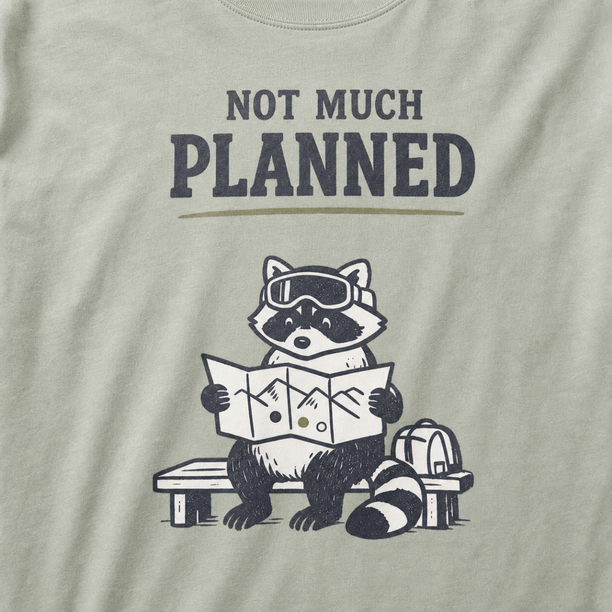Close-up of a khaki t-shirt featuring a raccoon in ski goggles holding a mountain map beside a backpack, with the words “Not Much Planned.”