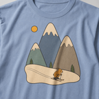 Close-up of a granite gray t-shirt featuring three muted snow-capped mountains, pine trees, a golden sun and a tiny animal skiing downhill.